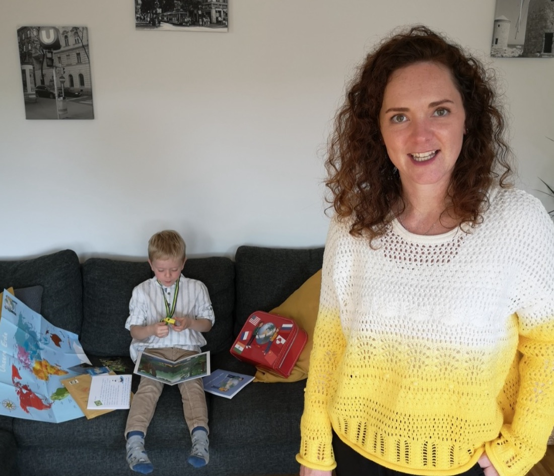 Mutter im Wohnzimmer, Sohn sitzt auf der Couch und liest in einem Länderbrief, daneben Koffer mit Länderflaggen